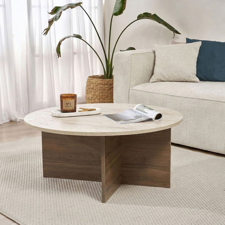 Runder Couchtisch mit Travertinplatte und dunklem Holzsockel in Kreuzform. Bank und Pflanze im Hintergrund.